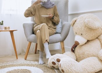 A women sitting on the chair sipping tea with a teddy bear placed beside her on the ground