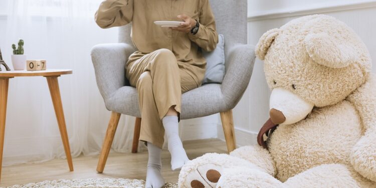 A women sitting on the chair sipping tea with a teddy bear placed beside her on the ground