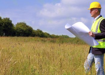 Architect wearing site safety gear and holding plans surveying a new building plot