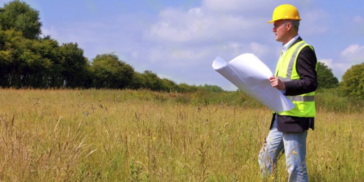 Architect wearing site safety gear and holding plans surveying a new building plot